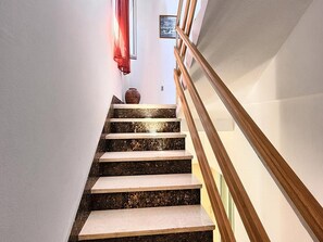 Una escalera con peldaños de colores alternos, claros y oscuros, barandal de madera y un jarrón en la plataforma.