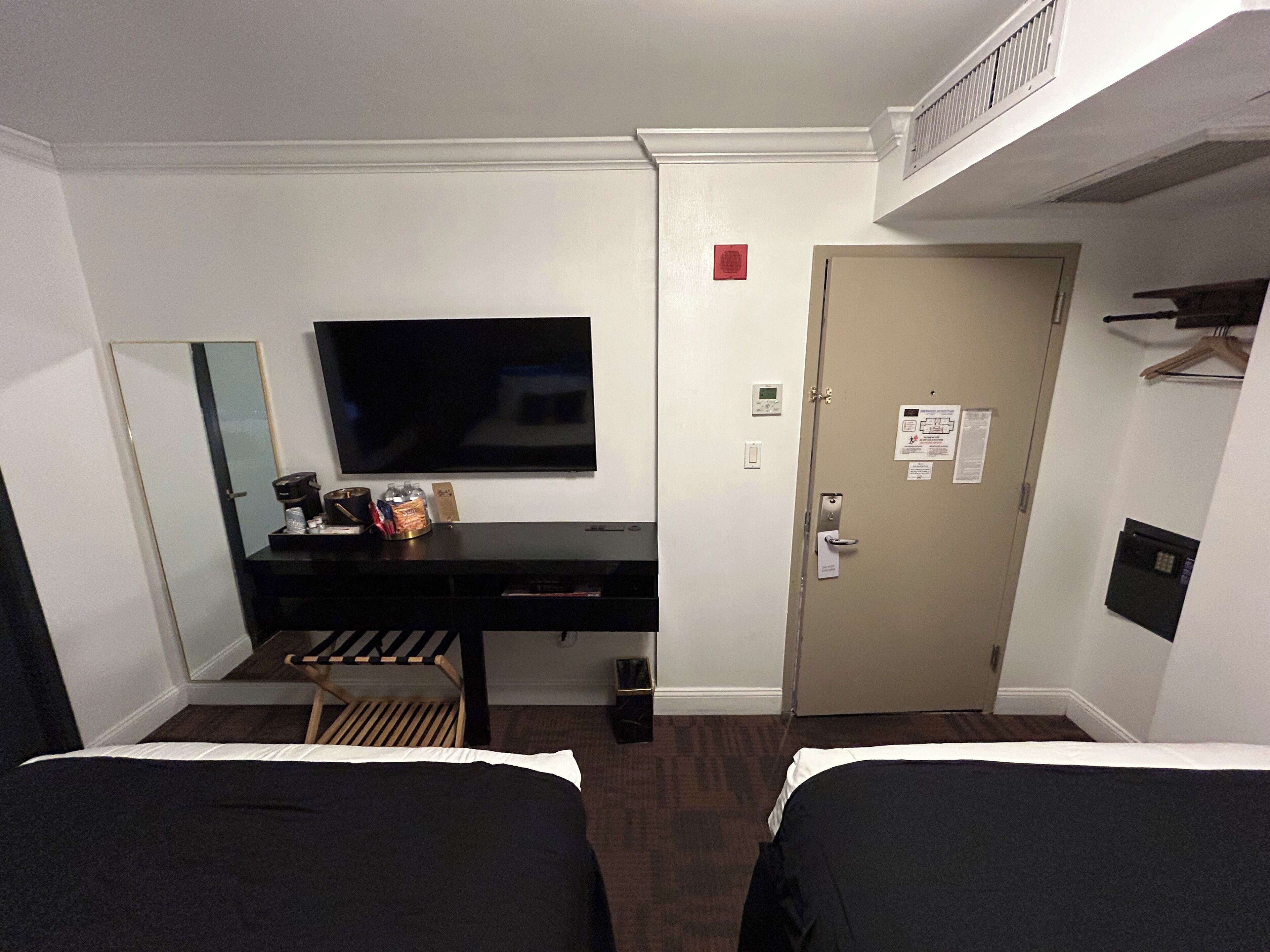 Une chambre d’hôtel avec deux lits, un bureau doté d’un téléviseur à écran plat, un miroir et une porte munie d’une serrure et d’un petit panneau rouge.