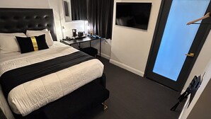 Ett modernt hotellrum med en stor säng, ett skrivbord med en lampa, en spegel och utsikt över himlen genom en dörr.