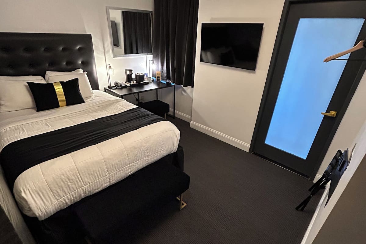 Ett modernt hotellrum med en stor säng, ett skrivbord med en lampa, en spegel och utsikt över himlen genom en dörr.