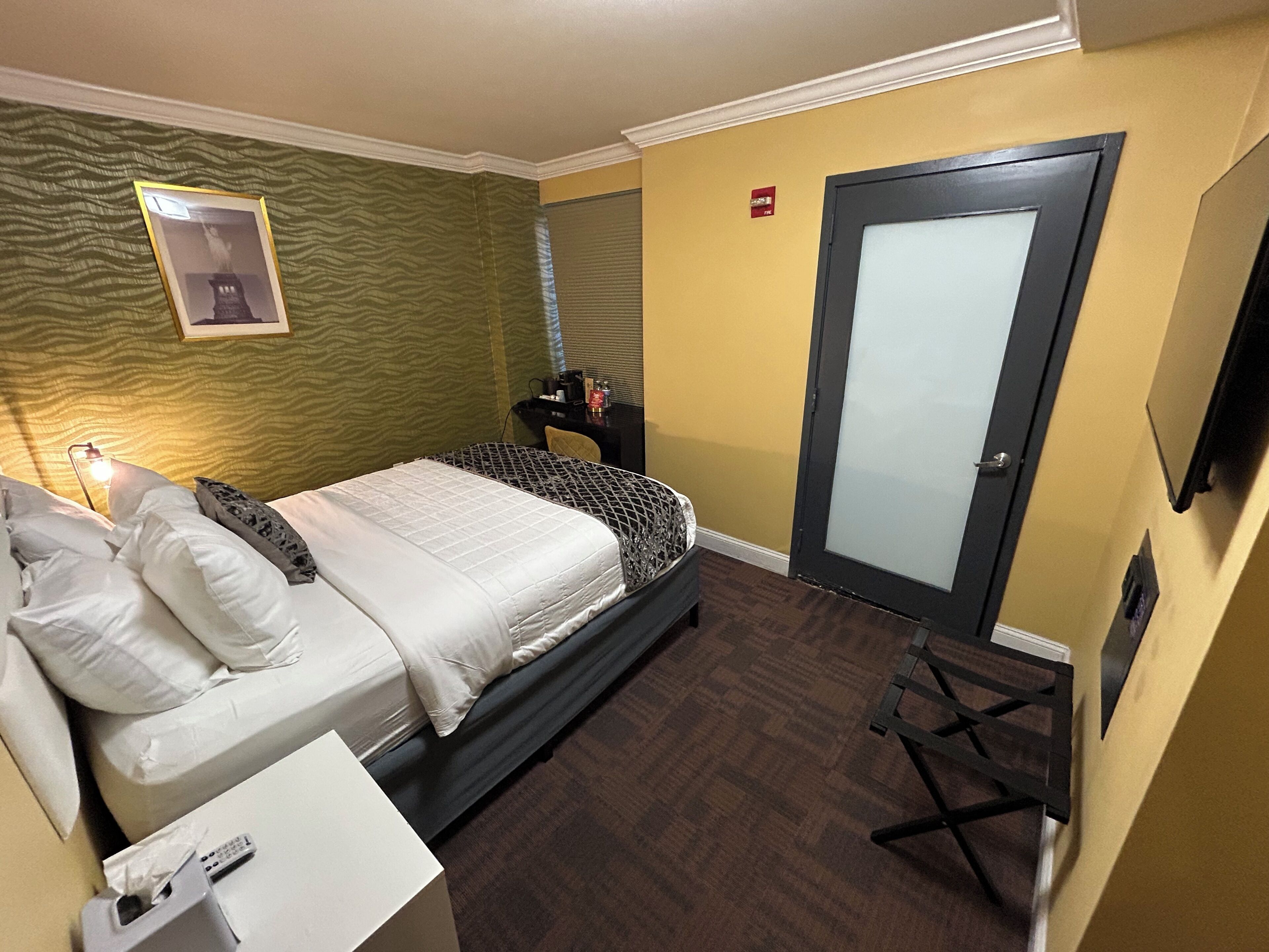 Una habitación de hotel con cama, mesita de noche y escritorio con silla. Se observa una puerta con panel de vidrio esmerilado y una pequeña señal roja encima.