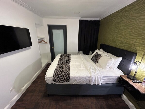 Habitación de hotel con cama, televisor montado en la pared, puerta y mesita de noche con lámpara y control remoto.