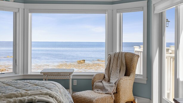 Ein Zimmer mit großem Erkerfenster mit Blick auf einen Strand, ein Korbstuhl und ein Bett mit gesteppter Decke.