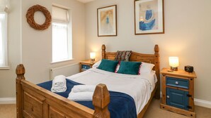 A neatly arranged bedroom with a wooden bed, two bedside tables with lamps, a window with a white blind, and two framed artworks on the wall. - Globe Cottage (Weymouth)