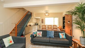 A spacious living room with a grey sofa, a wooden dining table, and a staircase leading to the kitchen. - Globe Cottage (Weymouth)