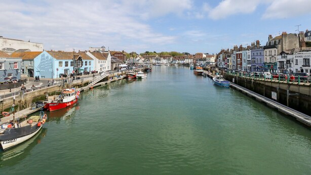 A harbor with boats docked along the waterfront, buildings lining the river, and a clear sky above. - Globe Cottage (Weymouth)