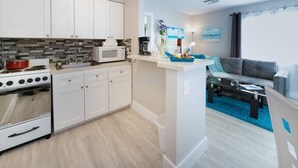 A modern kitchen with white cabinets, a microwave, and a stove. There is a dining area with a blue rug and a gray sofa. - Sea Star Suites By The Sea #6 Steps 2 Beach & Pubs (Lauderdale-by-the-Sea)