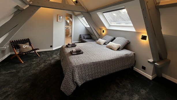 Un dormitorio con techo inclinado, una cama con ropa de cama estampada, una silla con cojín y un tragaluz.