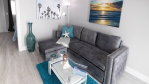 Un salon avec un canapé gris, une table basse en verre, un tapis turquoise et des œuvres d’art accrochées aux murs.