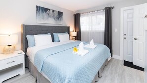 A neatly arranged hotel room with a bed, bedside tables, a nightstand, a headboard, a painting, a window with curtains, and a door. - Sea Star Suites By The Sea #7 Steps 2 Beach & Pubs (Lauderdale-by-the-Sea)