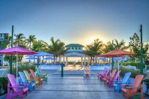 A resort area with colorful Adirondack chairs and umbrellas, a covered seating area, and palm trees.