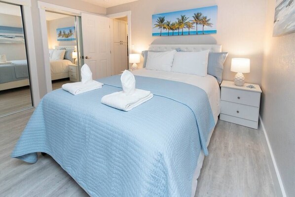 A hotel room with a large bed, a nightstand with a lamp, and a painting of palm trees on the wall.