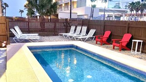 Una piscina con agua azul cristalina, rodeada de tumbonas, una cerca de madera y un edificio al fondo.