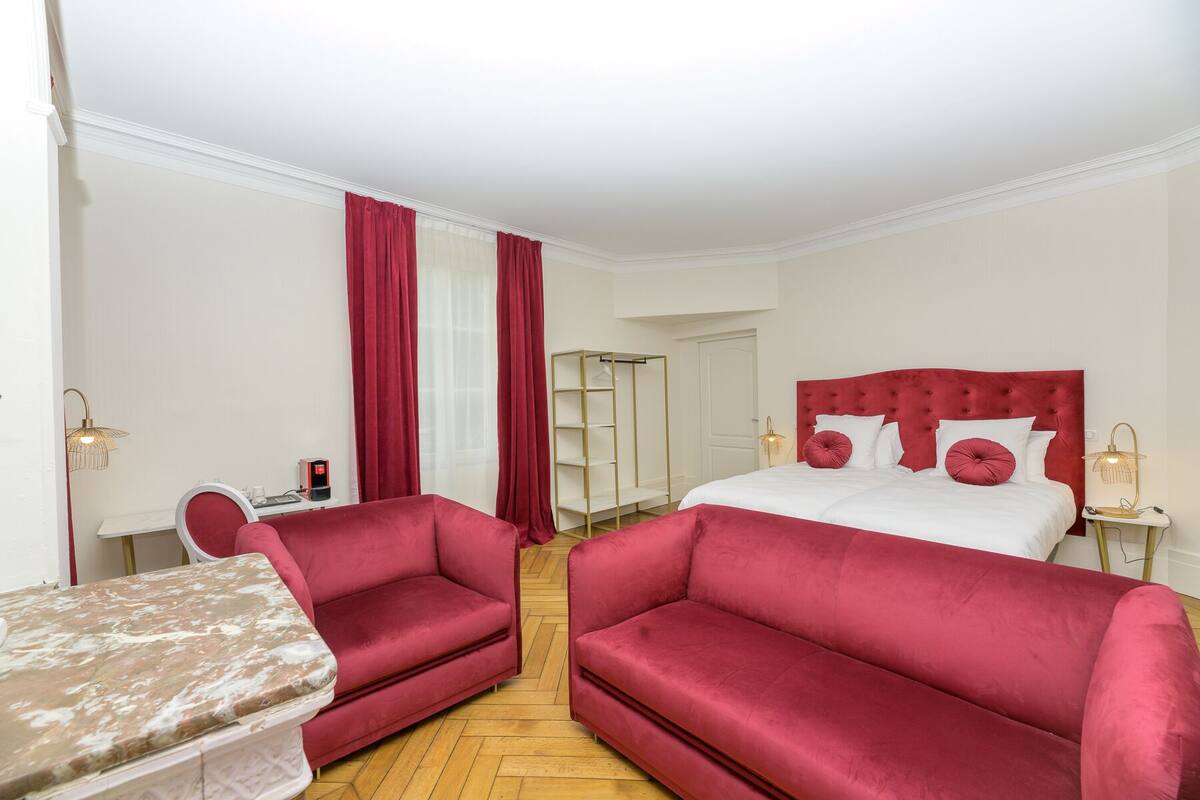 A hotel room with a large bed, two red sofas, a small table, and a mirror.
