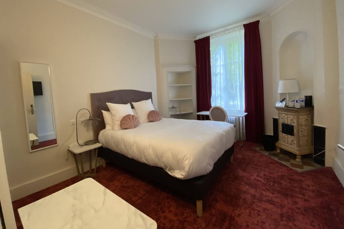 A hotel room with a large bed, a desk, a chair, a small table, a mirror, and a fireplace.
