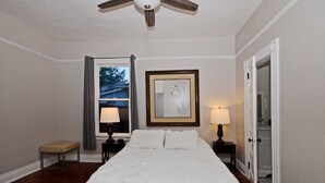 A bedroom with a bed, two bedside tables with lamps, a framed artwork on the wall, a window with curtains, and a door leading to a bathroom.