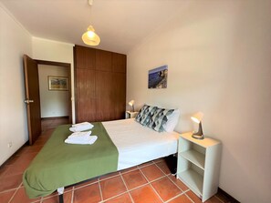 A bedroom with a bed, two bedside lamps, a green bedspread, a wooden wardrobe, and a painting on the wall.