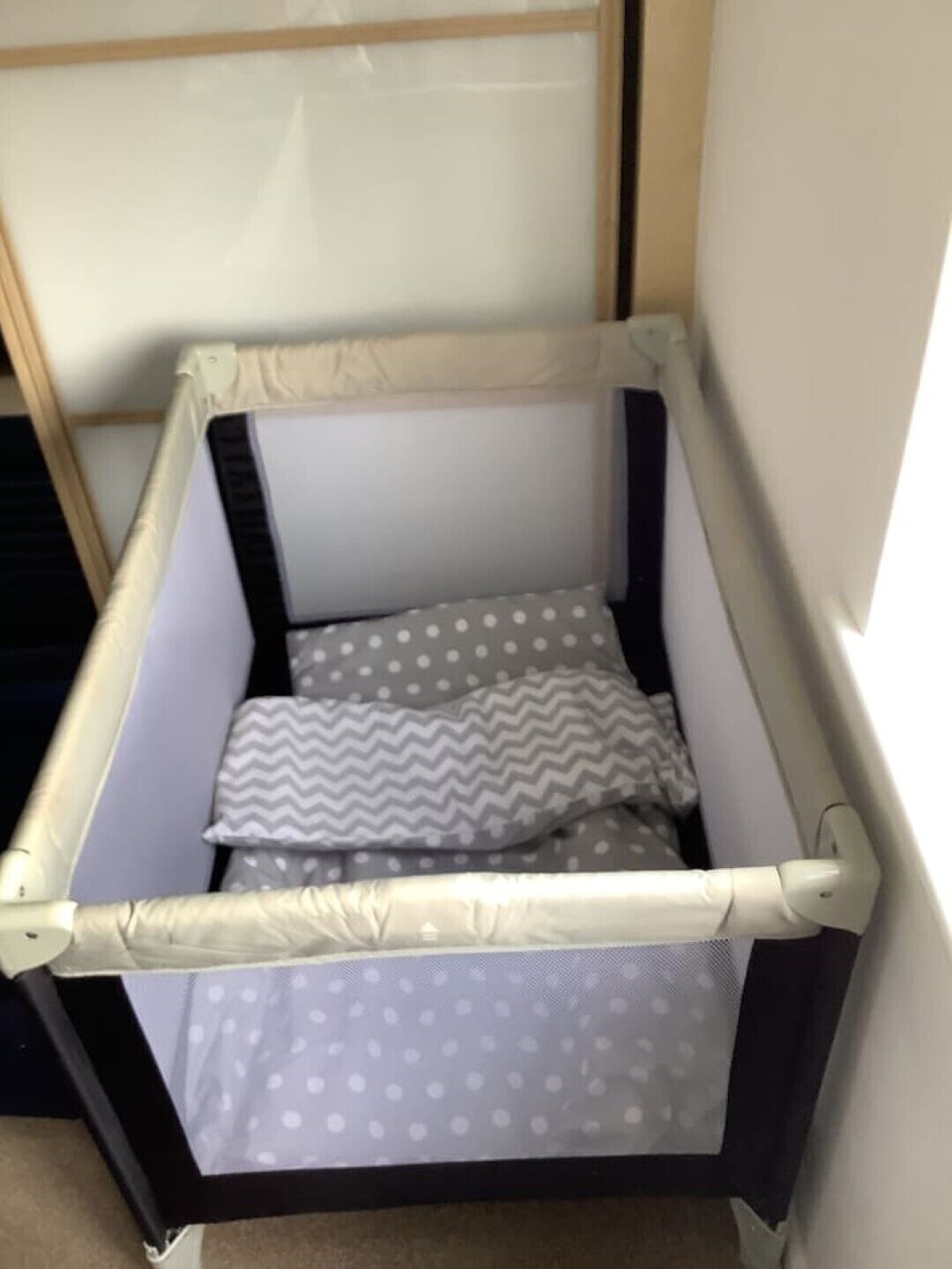 Una cuna con estructura blanca y negra, ropa de cama gris y blanca con lunares, y una almohada estampada en zigzag.