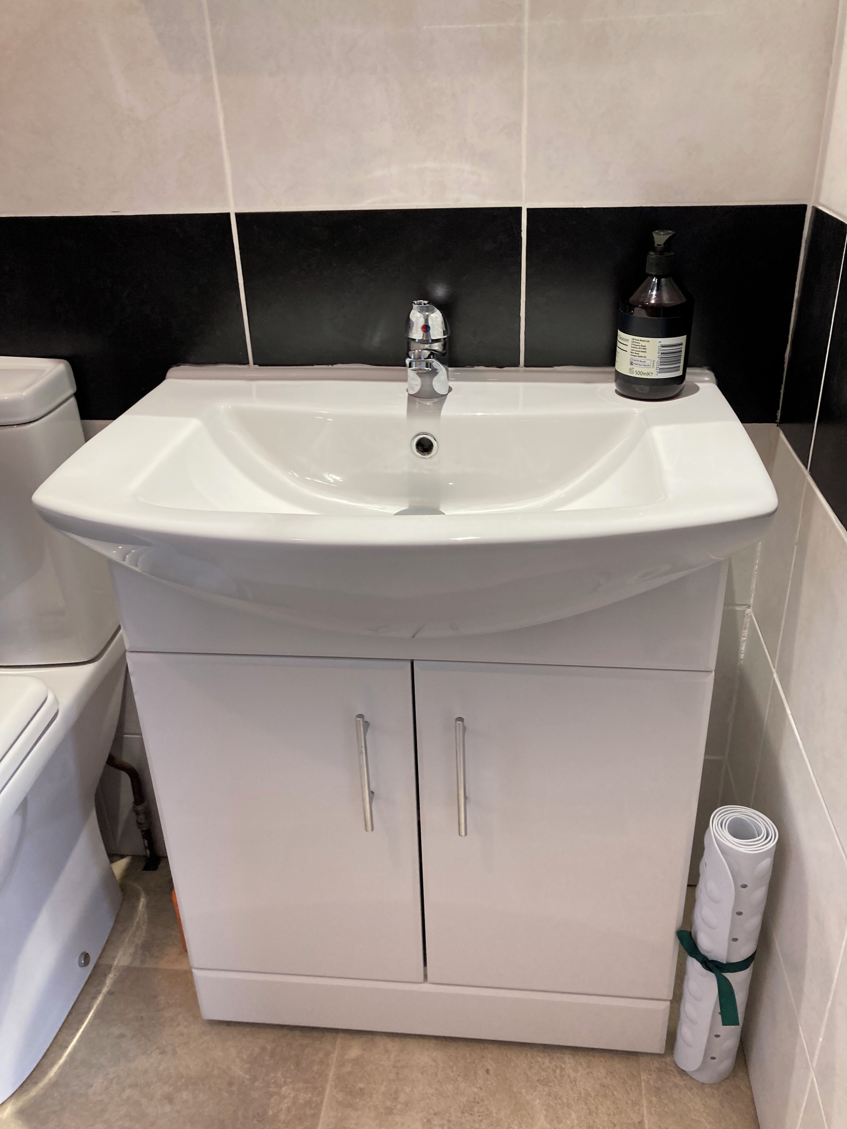 Un baño con un lavabo blanco, una botella negra de jabón para manos y una toalla blanca enrollada.