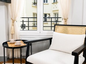 Una habitación con una silla de marco negro, una mesita con una bandeja y vistas a edificios a través de una ventana con barandillas negras.
