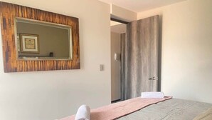 A neatly made bed with a pink towel and a rolled towel on a pink mat, a wooden-framed mirror on the wall, and a wooden door in the background. - Excelente Apto con Terraza Pegado a la Condesa (Ciudad de México)