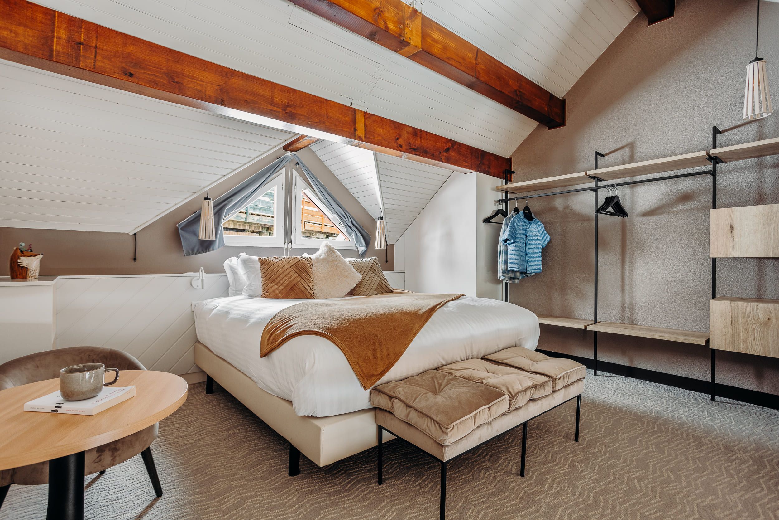 A bedroom with a large bed, a wooden ceiling, a window, a bedside table, and a hanging clothes rack.