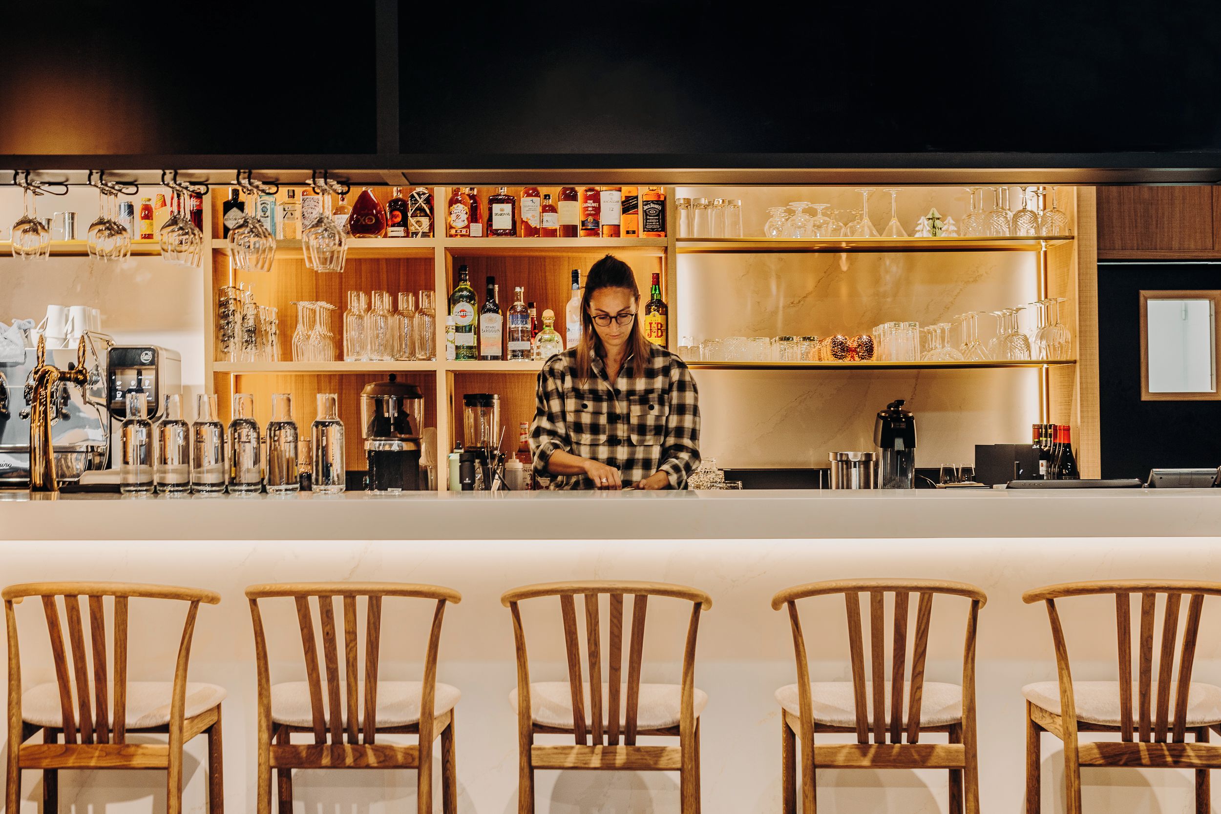 A bar area with wooden chairs, shelves with bottles, and a person working behind the bar.