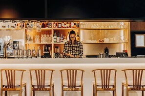 A bar area with wooden chairs, shelves with bottles, and a person working behind the bar. - Iskö Chalets-hötel (Eaux-Bonnes)