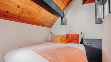 Une chambre avec un plafond en pente, un lit recouvert d’une couverture de couleur pêche, deux oreillers et une table de chevet avec une lampe.