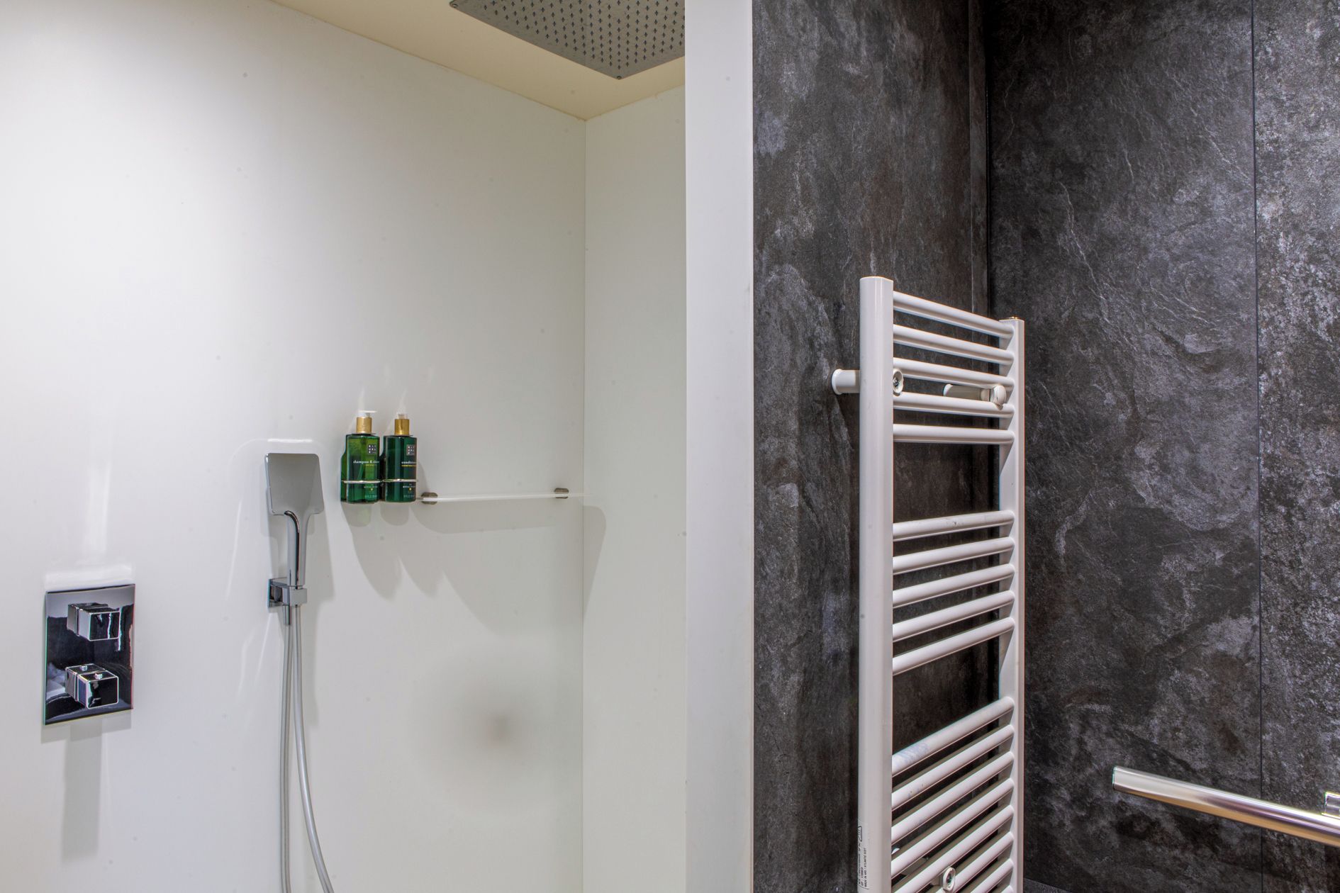 A bathroom with a white wall, a shower with a white towel rail, and two green bottles on the wall.