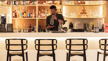 A bar area with a bartender preparing drinks, shelves stocked with various bottles, and three black chairs with woven seats.