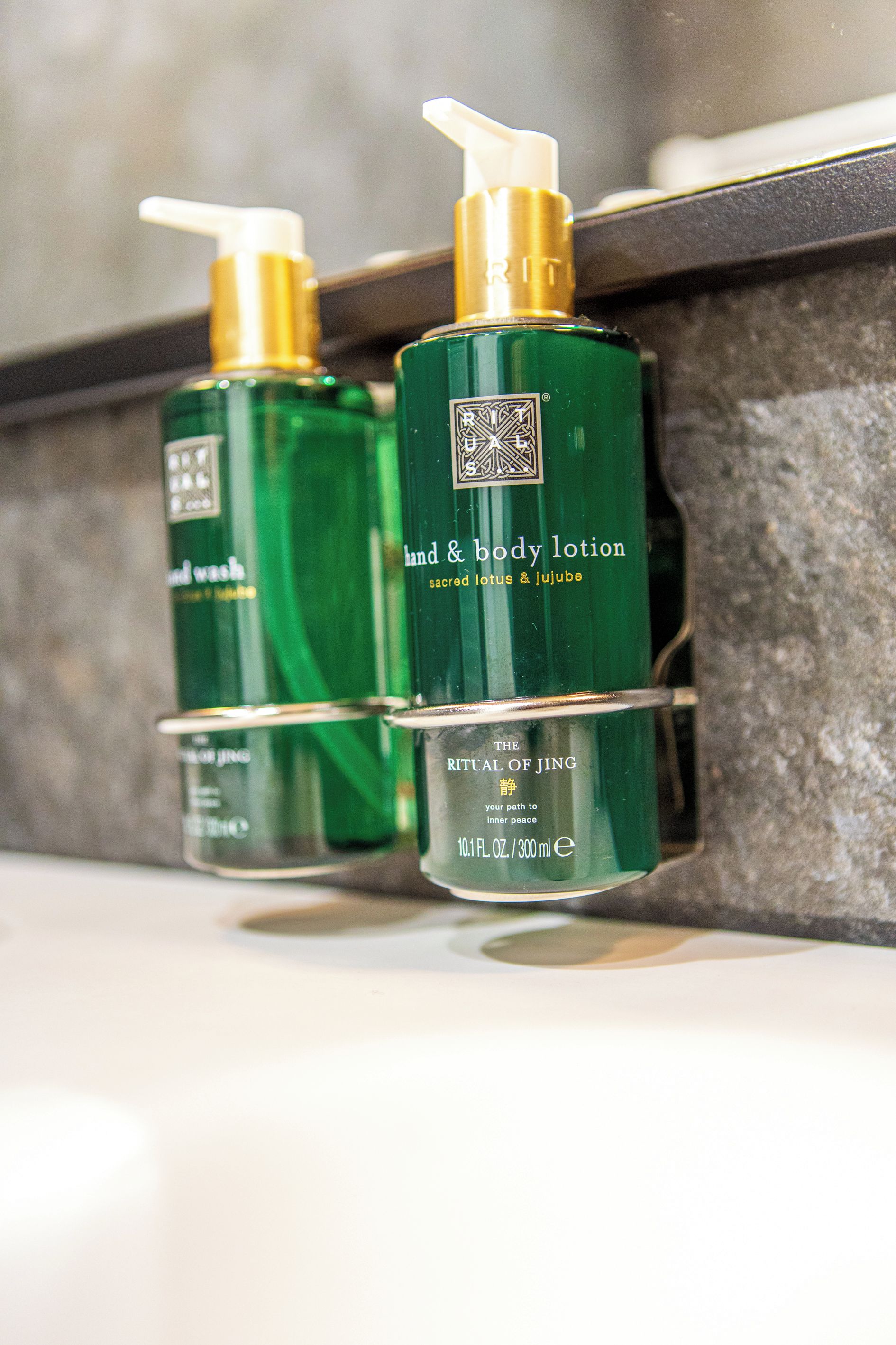 Zwei grüne Flaschen Hand- und Körperlotion mit Dosierern, beschriftet mit 'Sacred Lotus & Jujube' und 'The Ritual of Jing', stehen auf einem Regal.