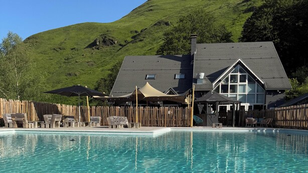 A clear swimming pool with lounge chairs and umbrellas, a wooden fence, and a building with a large window and a chimney in the background. - Iskö Chalets-hötel (Eaux-Bonnes)