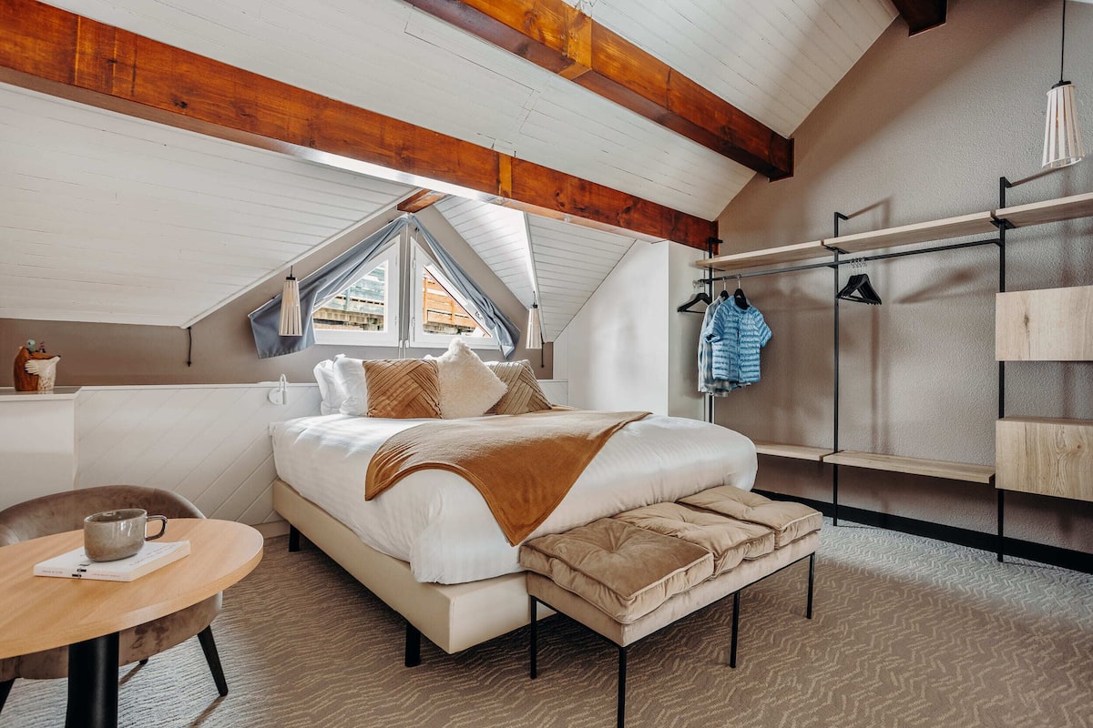 A bedroom with a large bed, a wooden ceiling, a window, a bedside table, and a hanging clothes rack.