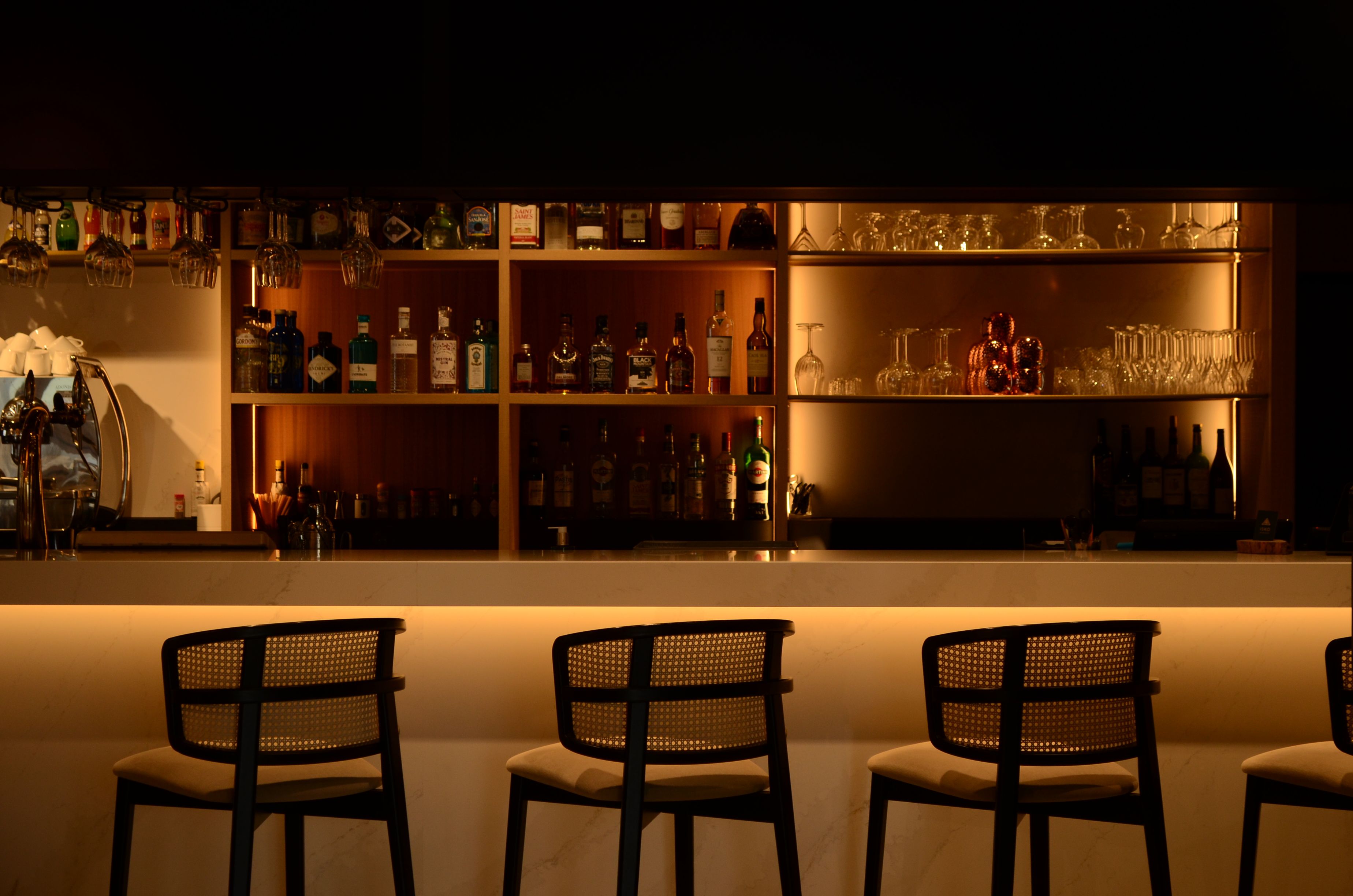 A bar area with shelves of liquor bottles and glasses, and three wicker chairs in the foreground.