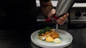 Eine Person mit Mütze gießt Soße aus einem Metallbehälter auf einen Teller mit Essen.