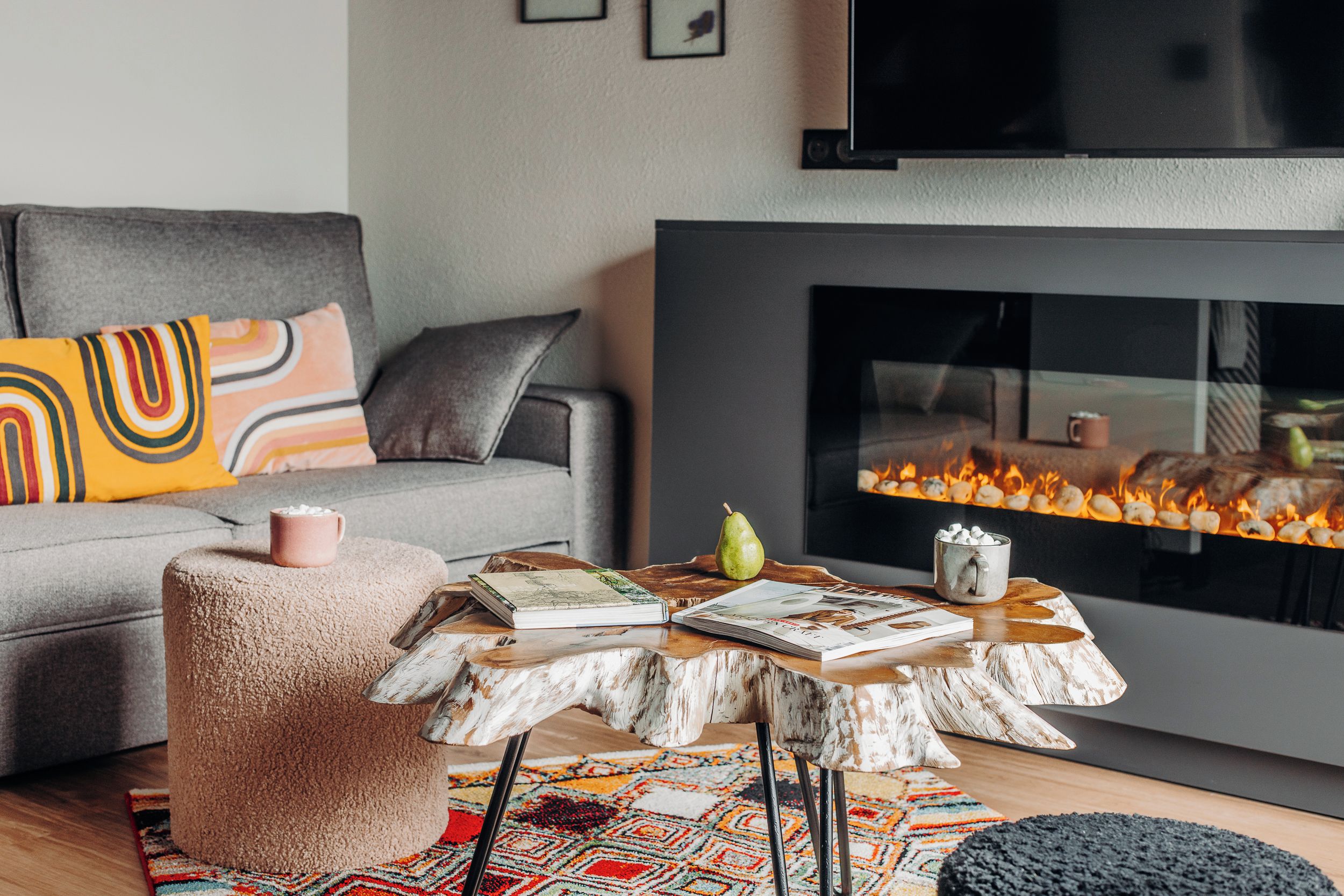 En moderne stue med en grå sofa, farverige puder, en rund skammel, et træsofabord med magasiner, en pære og en kop.