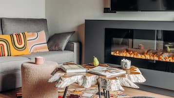 Uma sala de estar moderna com um sofá cinza, almofadas coloridas, um banquinho redondo, uma mesa de centro de madeira com revistas, uma pêra e uma caneca.