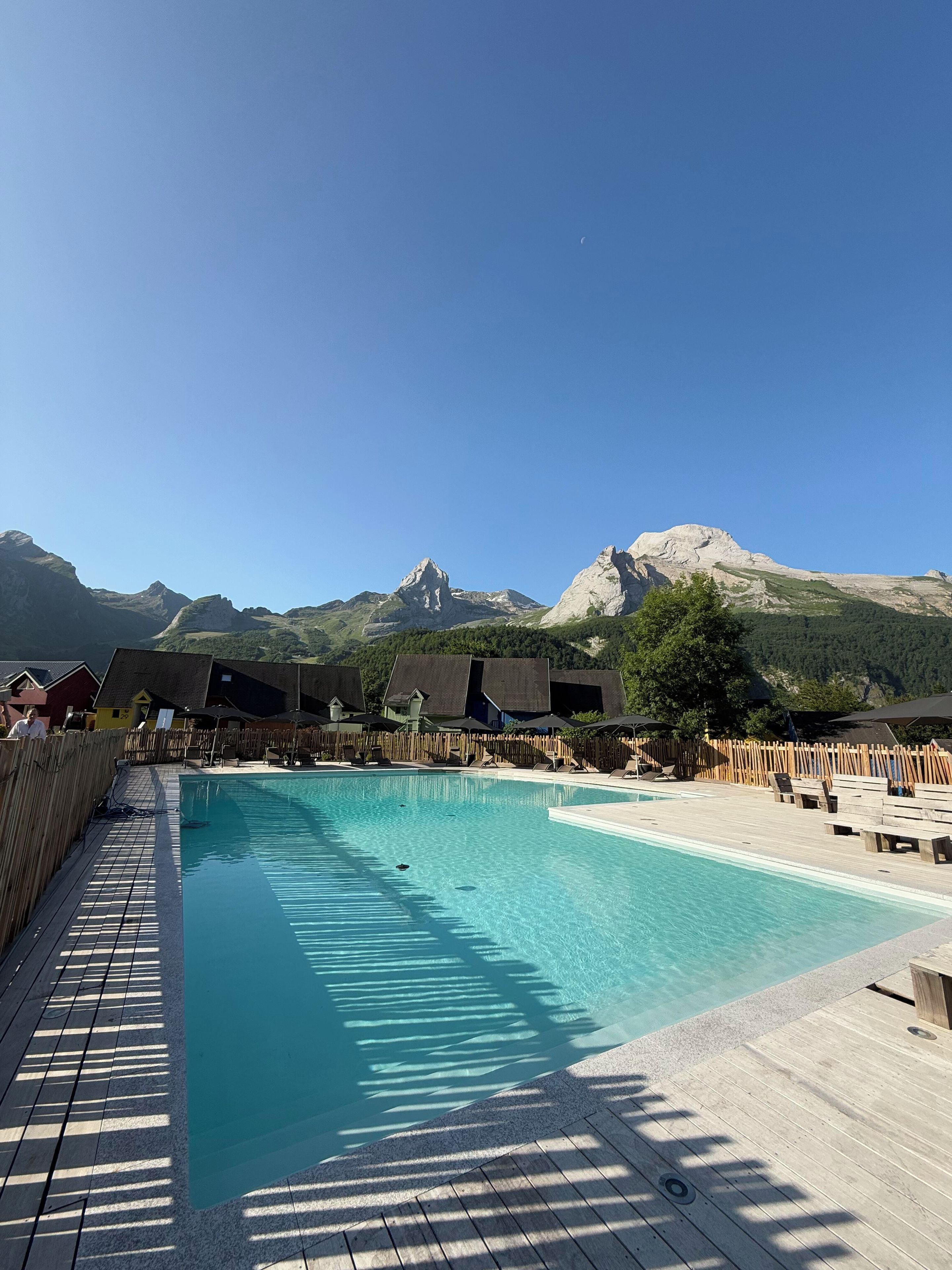 A swimming pool with wooden deck and lounge chairs, surrounded by mountains and chalet-style buildings.