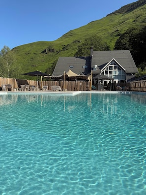 A clear swimming pool with lounge chairs and umbrellas, a wooden fence, and a building with a large window and a chimney in the background. - Iskö Chalets-hötel (Eaux-Bonnes)