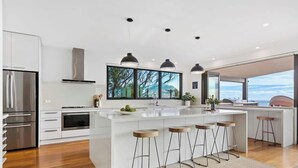 A modern kitchen with a large island, bar stools, stainless steel appliances, and a large window with a view. - The Ocean's Edge, Luxury Entertainer with Views (Mount Martha)