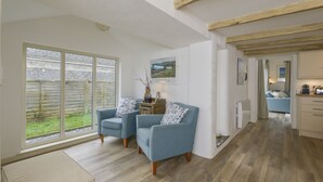 A spacious living area with two blue armchairs, a large window with a view of a garden, and a kitchen visible in the background. - Kelmscot (Port Isaac)
