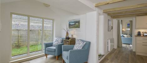 A spacious living area with two blue armchairs, a large window with a view of a garden, and a kitchen visible in the background.
