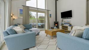 A living room with a sofa, coffee table, and fireplace. There is a TV mounted on the wall and a window with curtains. - Kelmscot (Port Isaac)