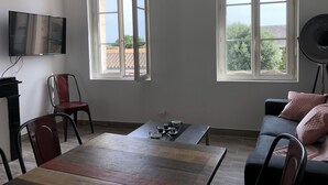Una habitación con una mesa de comedor, dos sillas, un sofá, un televisor y dos ventanas con contraventanas abiertas.