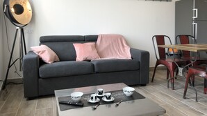 Una sala de estar con un sofá gris, una mesa de centro con un control remoto, tazas y un bol. Se observa un calendario en la pared y un refrigerador al fondo.