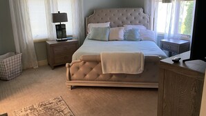 Une chambre à coucher avec un grand lit, deux tables de chevet, un ventilateur de plafond et une fenêtre avec des rideaux.