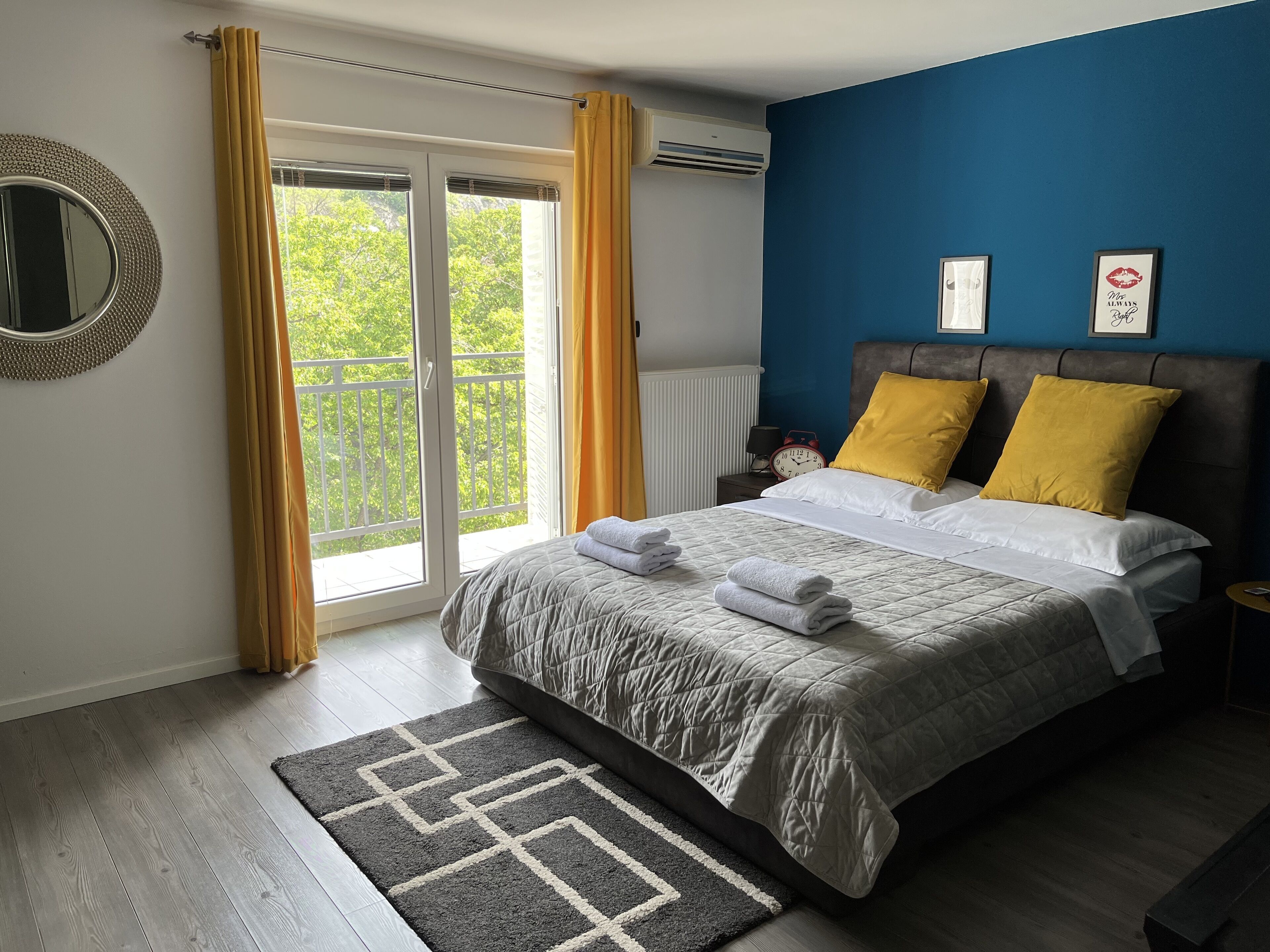 Un dormitorio con cama, mesita de noche, reloj y vistas a la vegetación por la ventana.