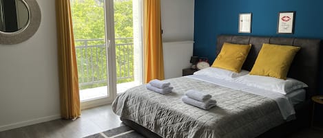 A bedroom with a bed, a nightstand, a clock, and a view of greenery through the window.