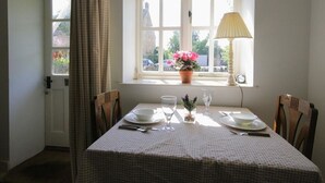 A dining area with a table set for two, a window with a view of the outside, a lamp on the windowsill, and a door with a curtain. - Noel Cottage (Chipping Campden)